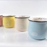 Four pastel-colored mugsโpink, yellow, cream, and blueโare arranged in a line on a white surface, each with a different word and animal illustration on the front.