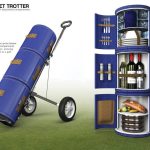 A blue, cylindrical, wheeled picnic hamper stands upright and open, revealing compartments with tableware, wine bottles, glasses, plates, cutlery, and food items.