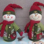 Two plush snowman dolls wear red hats, green jackets, and red scarves, standing side by side against a tiled background.