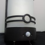 A cylindrical wall-mounted lamp with a frosted white shade, black metal accents, two pull chains, and a motion sensor on a black base.