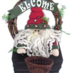 A festive Santa Claus doll with a long white beard sits in front of a basket, framed by a wreath with greenery and a sign reading "WELCOME.