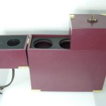 A burgundy wooden box with brass accents, featuring compartments, circular cutouts, a side drawer, and a leather strap.