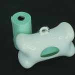 A bone-shaped white plastic dispenser and a roll of green dog waste bags are shown against a black background.
