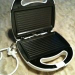 An electric sandwich maker with its lid open, showing the non-stick grilling plates and a white power cord with a plug.