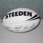 A white Steeden rugby ball with black lettering and multiple signatures, placed on a light grey surface against a textured background.