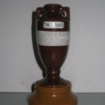 A replica of The Ashes trophy, featuring a dark brown urn with two handles and a label on a round, light brown base.