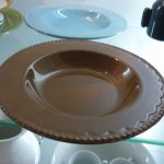 A brown ceramic soup plate with a scalloped edge is displayed on a glass shelf among other dishes and tableware.