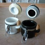Two small ceramic canisters, one white and one black, with metal clasps and miniature wooden spoons attached, are open and placed side by side on a wooden surface.