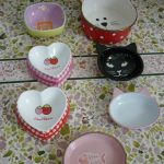 Assorted ceramic bowls, some heart-shaped and cat-themed, arranged on a tiled surface. Designs include strawberries, a fish, and cat faces with ears.