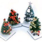 Four small decorated Christmas trees, each on a white base, adorned with ornaments, ribbons, and figurines, including Santa Claus and a snowman.