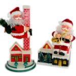 Two Santa Claus figurines: one climbing a red brick chimney with a green sack, the other sitting on a rooftop playing a gold saxophone. Both are surrounded by small model houses.