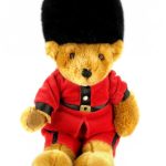 A teddy bear dressed as a British royal guard, wearing a red uniform and a tall black hat, sits against a white background.