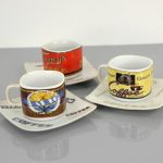 Three ceramic espresso cups with matching saucers, each featuring different vintage coffee-themed designs, are arranged on a grey surface.