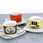 Three ceramic coffee cups with different vintage designs are placed on square white saucers with the word "COFFEE" printed on them, set on a gray surface.