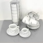 A set of white cups and saucers on a grey mat, with some cups stacked vertically in a metal holder and others hanging on a rack.