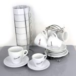 A set of white ceramic coffee cups with saucers, some stacked in metal racks and others on a table, labeled "Gimoka.