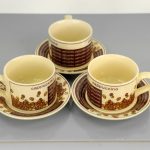 Three ceramic cappuccino cups with matching saucers, decorated with coffee bean and text designs, are arranged on a gray surface.