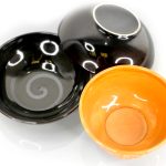 Three ceramic bowls are displayed: one black bowl with a swirl pattern, one plain orange bowl, and one black bowl turned upside down.