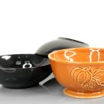 Three ceramic bowls: two plain black bowls and one orange bowl with a raised pumpkin design, arranged close together on a white surface.