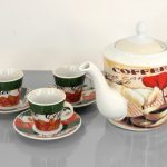 A ceramic teapot and three matching cups with saucers, all decorated with coffee-themed designs, displayed on a gray surface.