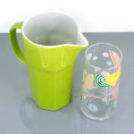 A green ceramic pitcher is placed next to a transparent glass decorated with colorful circular patterns on a gray surface.