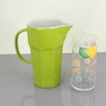 A green ceramic pitcher is placed next to a clear glass with colorful circular patterns on a gray surface.