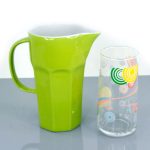 A green ceramic pitcher with a handle is placed beside a clear glass decorated with colorful circular patterns on a gray surface.