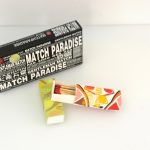 A black box labeled "MATCH PARADISE" and two smaller matchboxes, one open showing wooden matches with red tips, are placed on a white surface.