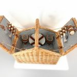 A wicker picnic basket with a striped interior, open to show plates, cups, and cutlery secured inside the lid with leather straps.