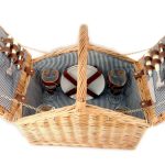 Open wicker picnic basket with striped lining, containing plates, cutlery, wine glasses, and bottle holders secured with leather straps.