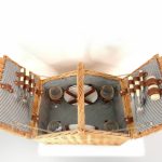 Open wicker picnic basket with striped fabric lining, containing plates, cups, cutlery, and salt and pepper shakers neatly secured in leather straps.