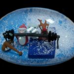 A snow globe with Santa, a snowman, and a reindeer lifting a barbell together, set on a blue platform with artificial snow inside.