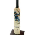 A cricket bat featuring a printed image and signature of Sachin Tendulkar is mounted on a wooden base with a calendar and nameplate.