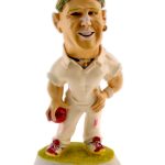A caricature figurine of a cricket player in white uniform holding a red ball, standing on a base labeled "SHANE WARNE.