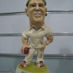 A caricature figurine of a cricket player holding a ball, standing on a base labeled "SHANE WARNE," against a background with horizontal metallic lines.