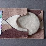 Two matching brown and beige bathroom rugs are displayed on a grey carpet, one rectangular and one U-shaped, with a Suya brand label attached.