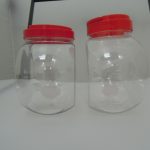 Two empty clear plastic jars with red lids are placed side by side on a white surface, with a black bar in the background.