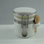 A small white ceramic jar with a metal clasp and lid, featuring an attached spoon and a small handle, placed on a white surface against a gray background.